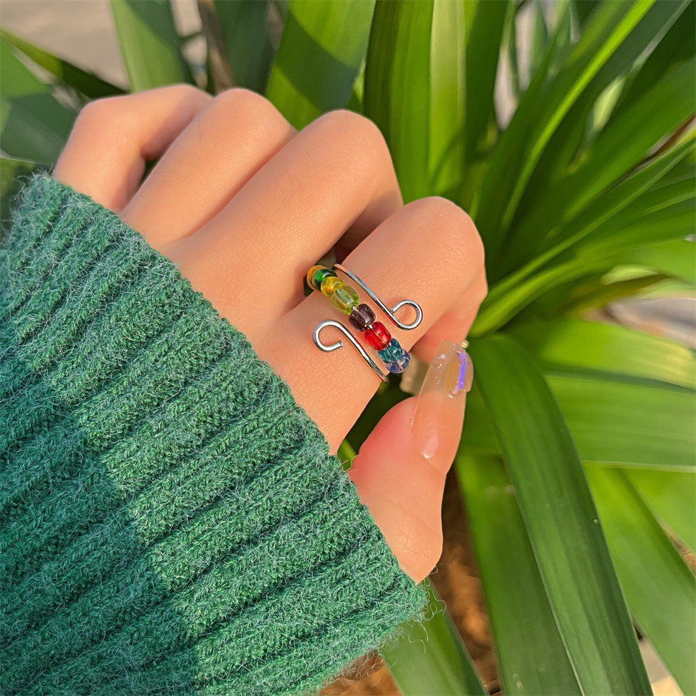 A hand in a green knit sweater displays the Adjustable Beaded Crochet & Anxiety Relief Tool ring, featuring colorful beads, with green leafy plants in the background.