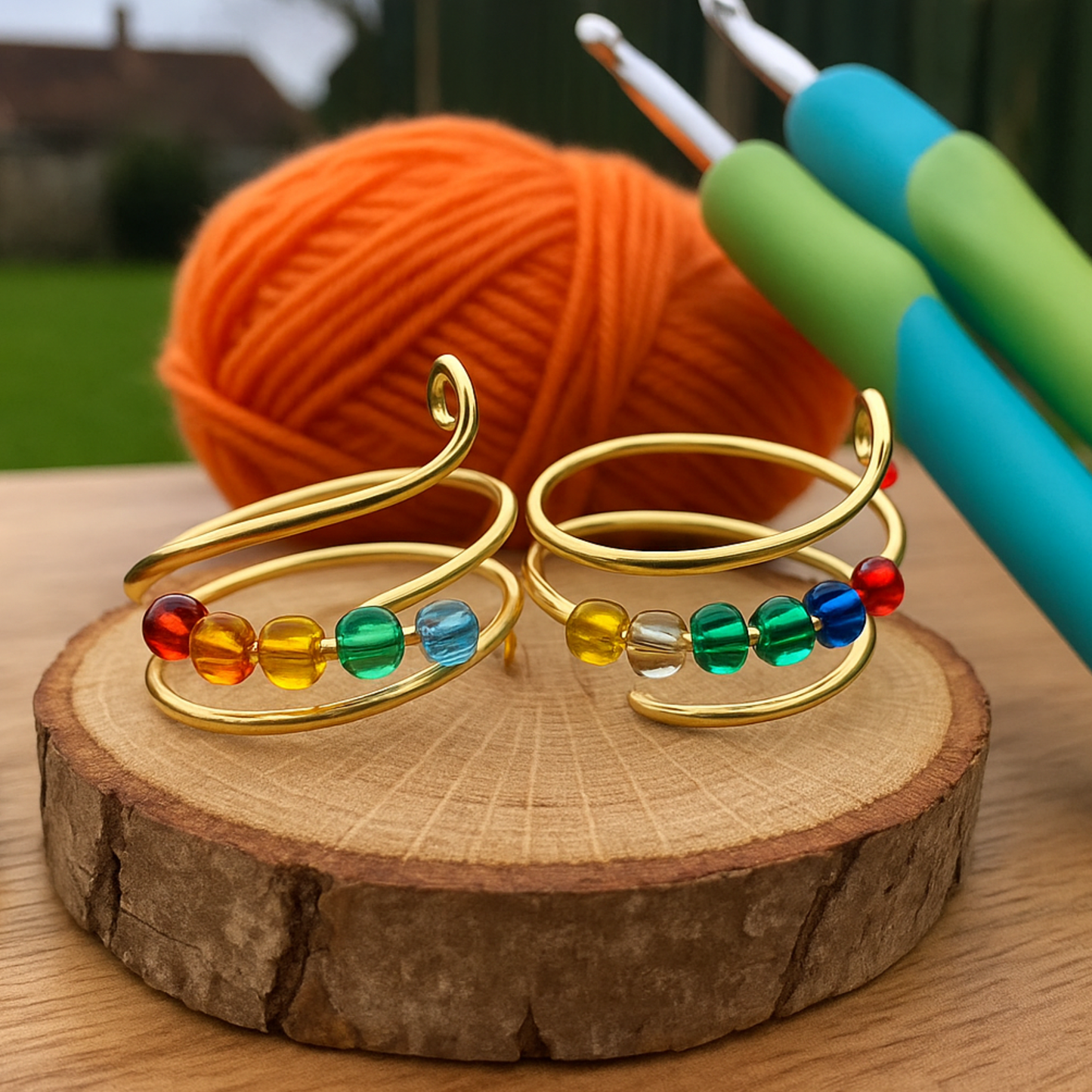 The Adjustable Beaded Crochet & Anxiety Relief Tool features gold wire tension rings with rainbow beads on a wooden slice, accompanied by two crochet hooks with blue and green handles, plus an orange yarn ball, set outdoors with a blurred background.