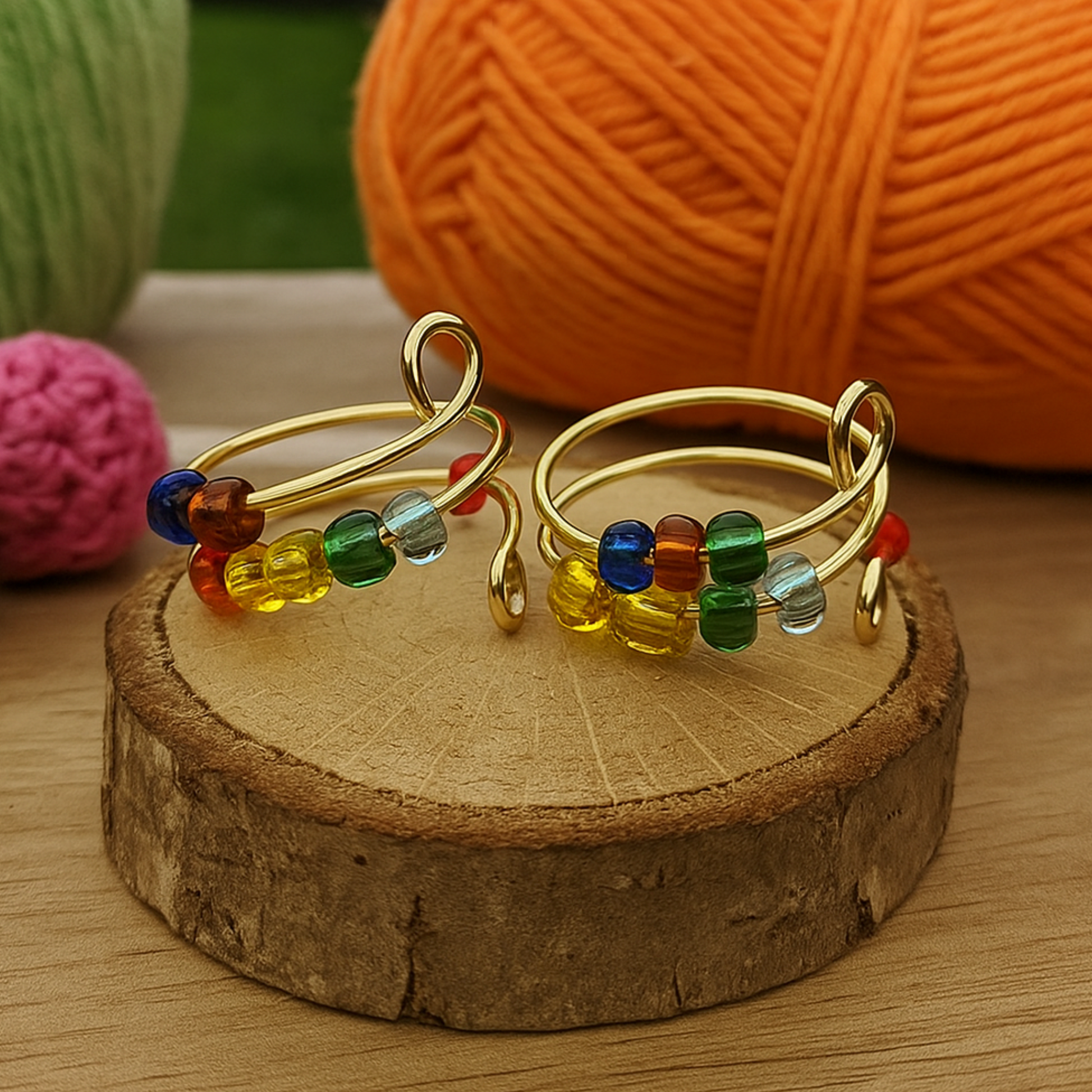 The Adjustable Beaded Crochet & Anxiety Relief Tool features two gold wire rings with colorful glass beads, shown on a wooden slice. Orange, green, and pink yarn balls sit in the background on a wooden surface.