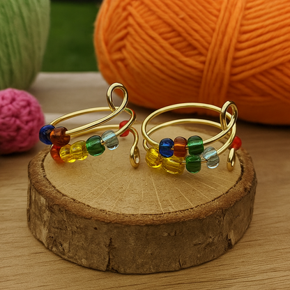 The Adjustable Beaded Crochet & Anxiety Relief Tool features two gold wire rings with colorful glass beads, shown on a wooden slice. Orange, green, and pink yarn balls sit in the background on a wooden surface.